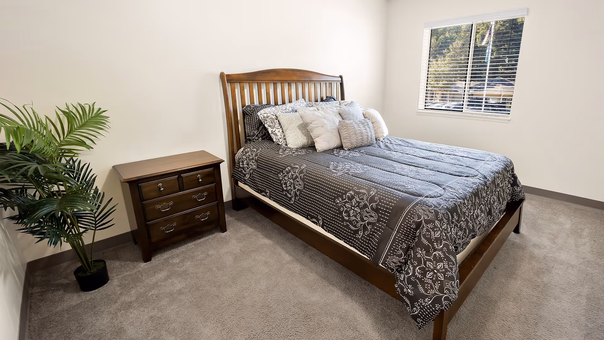 A bedroom with a wooden bed frame and a bed made with a black and white floral patterned comforter and multiple pillows. There is a wooden nightstand with drawers next to the bed and a potted plant in the corner. A window with white blinds lets in natural light.