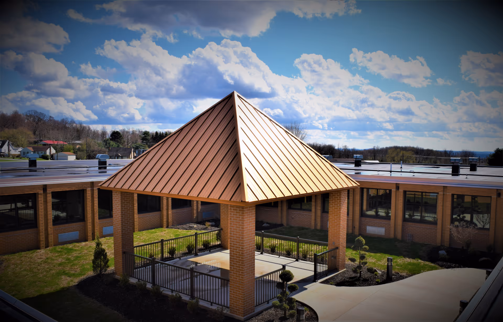 Outdoor courtyard area of a senior living facility featuring a covered pavilion with a copper-colored metal roof supported by brick columns, surrounded by a black metal fence. The courtyard is enclosed by a single-story brick building with large windows. The sky is partly cloudy with blue patches visible.