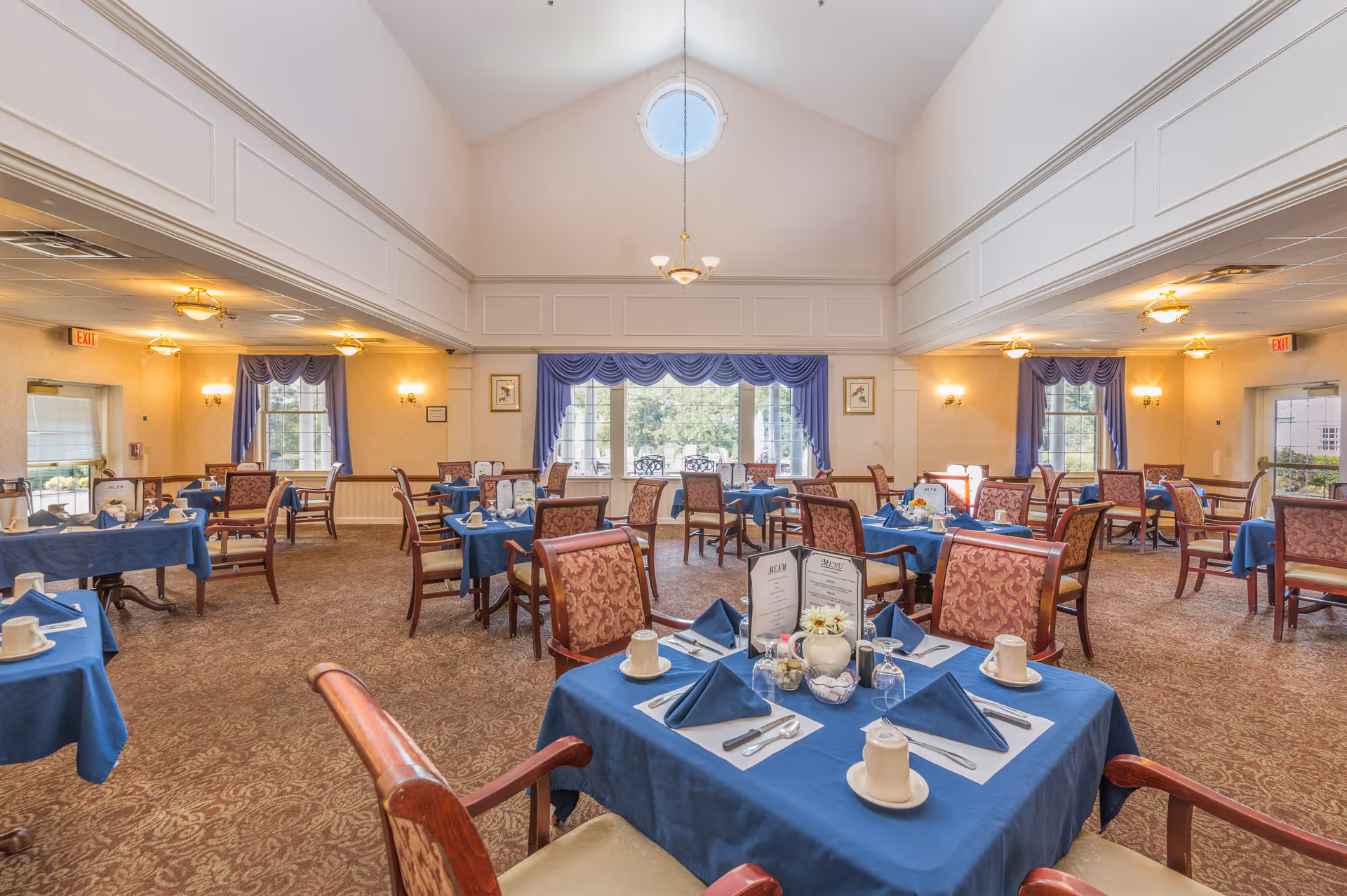 A spacious dining room with multiple tables covered in blue tablecloths, each set with white cups, silverware, and folded blue napkins. The room features high ceilings with a round window near the top, large windows with blue curtains, patterned carpet, and upholstered chairs with wooden frames. Soft lighting fixtures hang from the ceiling and walls.