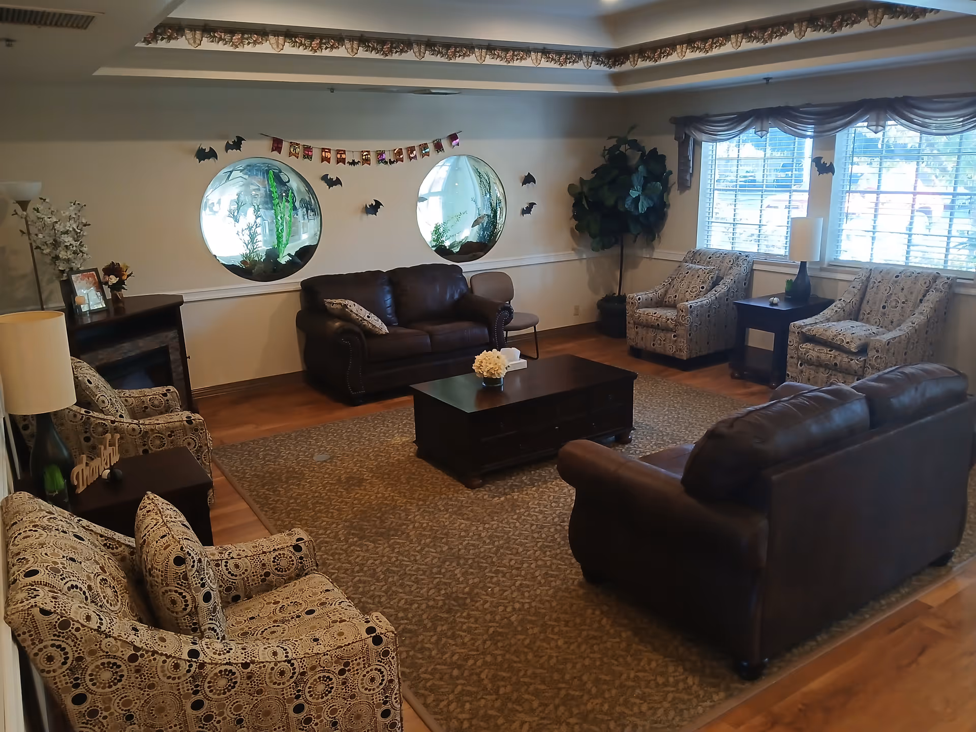 A cozy living room with two brown leather sofas, four patterned armchairs, a dark wooden coffee table, and a side table with a lamp. The room has hardwood floors and a large area rug. Two round windows with fish tanks are set into the wall, decorated with small bat cutouts and a Halloween banner. A large potted plant is in the corner near windows with blinds.