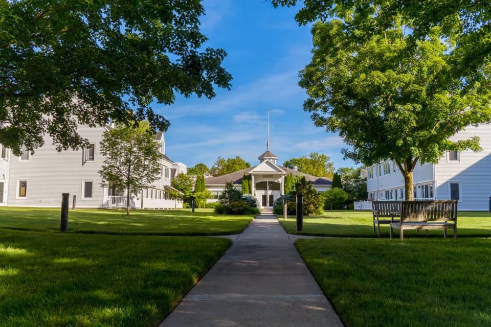 A paved walkway leads through a well-maintained green lawn with trees and benches on either side, flanked by two white multi-story buildings, under a clear blue sky.