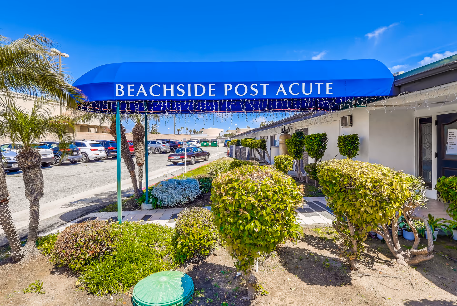 Entrance to Beachside Post Acute facility with a blue awning displaying the facility name. The entrance is surrounded by trimmed bushes and palm trees, with a parking lot and several cars visible in the background under a clear blue sky.