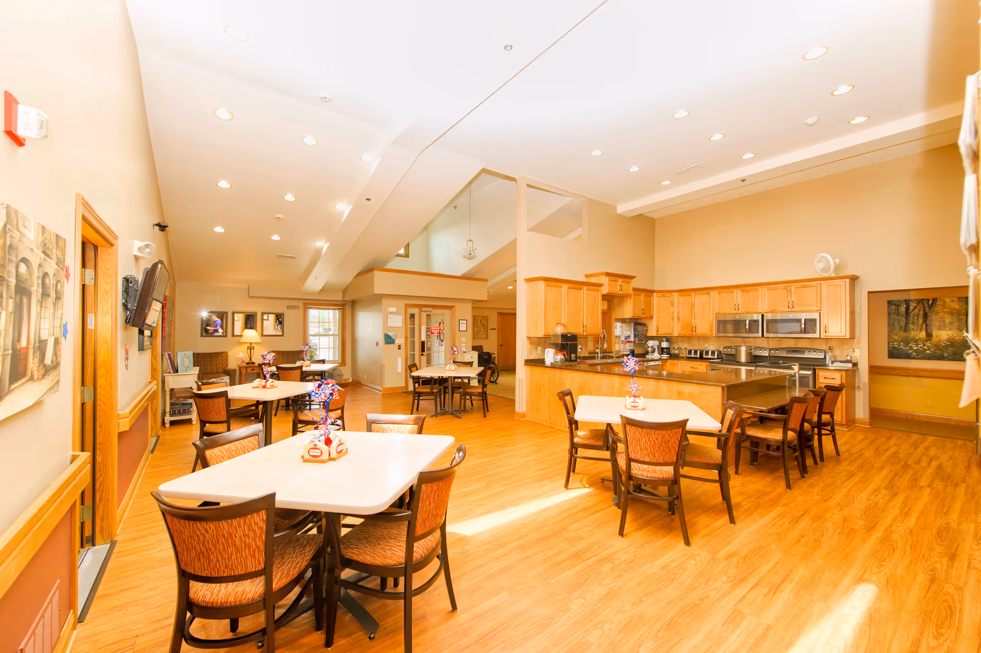 A spacious, well-lit dining area with multiple tables and chairs arranged on a wooden floor. The room features a kitchen area with wooden cabinets, stainless steel appliances, and a countertop. The walls are painted beige, and there are decorative items on the tables and walls. The ceiling has recessed lighting, and there is a window letting in natural light.