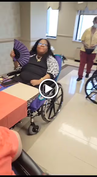 An elderly woman in a wheelchair holding a purple fan is seated at a table covered with a red tablecloth in a bright room with windows. Another person stands nearby, also near a wheelchair. The setting appears to be a communal indoor space.