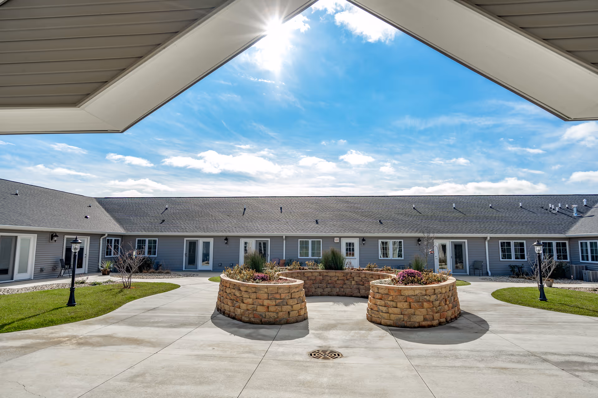 Outdoor courtyard area of Villas of Holly Brook Assisted Living & Memory Care in Pekin, IL, featuring a circular stone planter with flowers and shrubs, surrounded by a paved walkway and a single-story building with multiple doors and windows under a bright blue sky with some clouds.