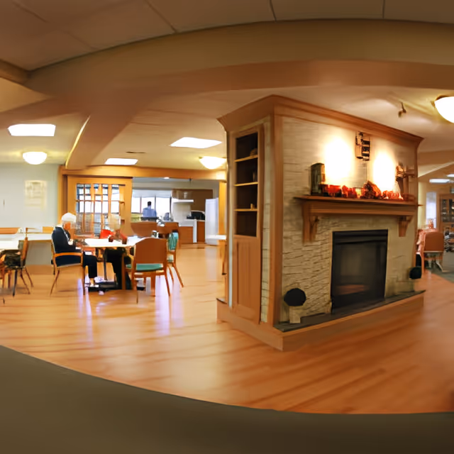 Interior view of a senior living facility common area with wooden flooring, a stone fireplace with a wooden mantle, and several tables and chairs. An elderly person is seated at one of the tables. The background shows a kitchen area and additional seating.