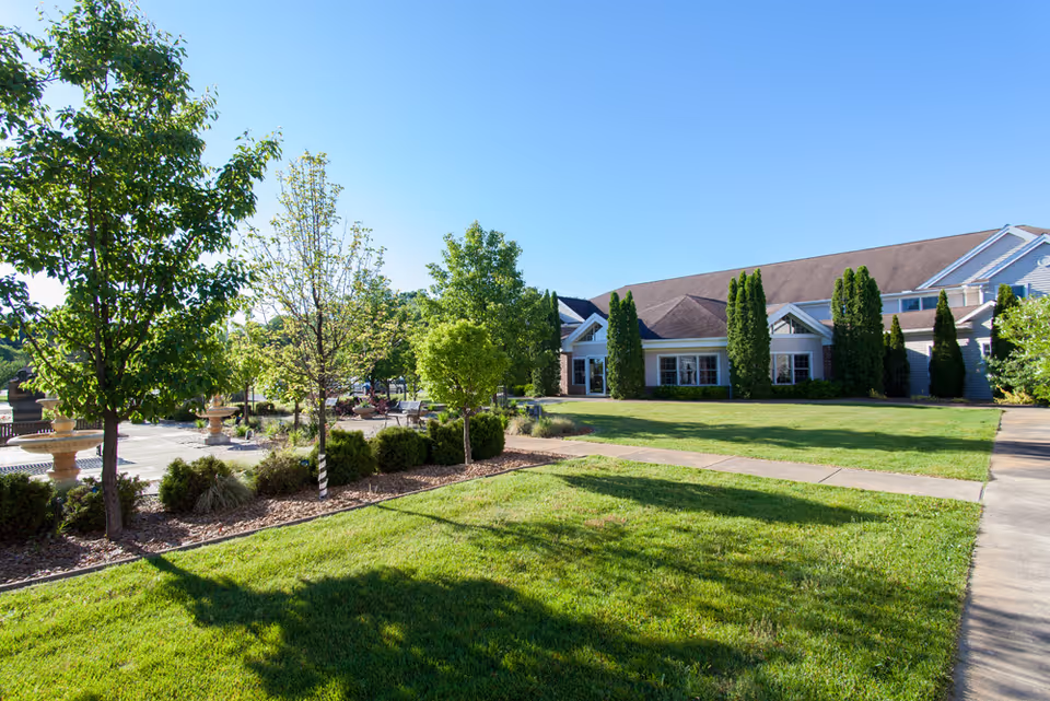 A sunny outdoor view of a retirement community with a well-maintained lawn, several trees, shrubs, and a paved walkway leading to a large building with multiple windows and a sloped roof.
