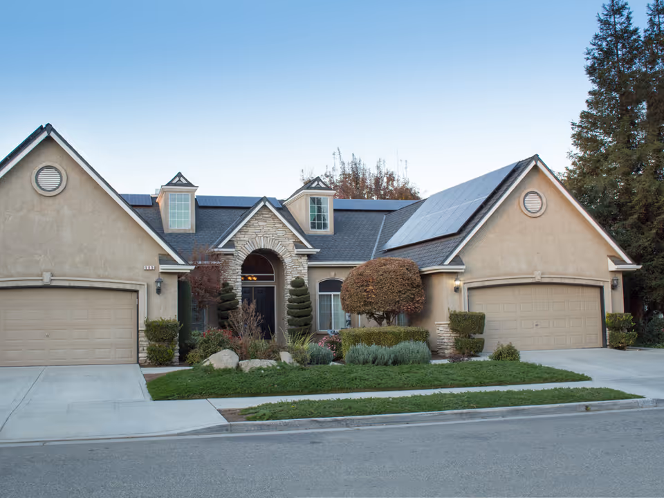 Front exterior view of a single-story residential building with two garages, a central entrance with stone archway, manicured shrubs, and solar panels on the roof under a clear sky.