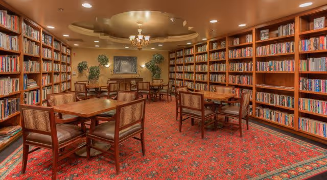 A cozy library room with wooden bookshelves filled with books lining the walls. Several wooden tables with cushioned chairs are arranged on a red patterned carpet. The ceiling has recessed lighting and a decorative chandelier, with some green plants and framed artwork on the far wall.