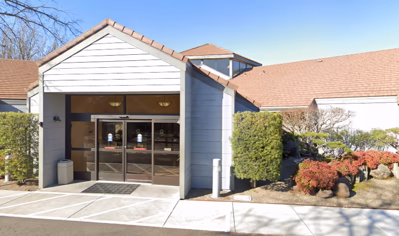 Exterior front entrance of ACC Care Center: Skilled Nursing & Rehabilitation with automatic sliding glass doors, a small covered entryway, and landscaped bushes and trees on either side.