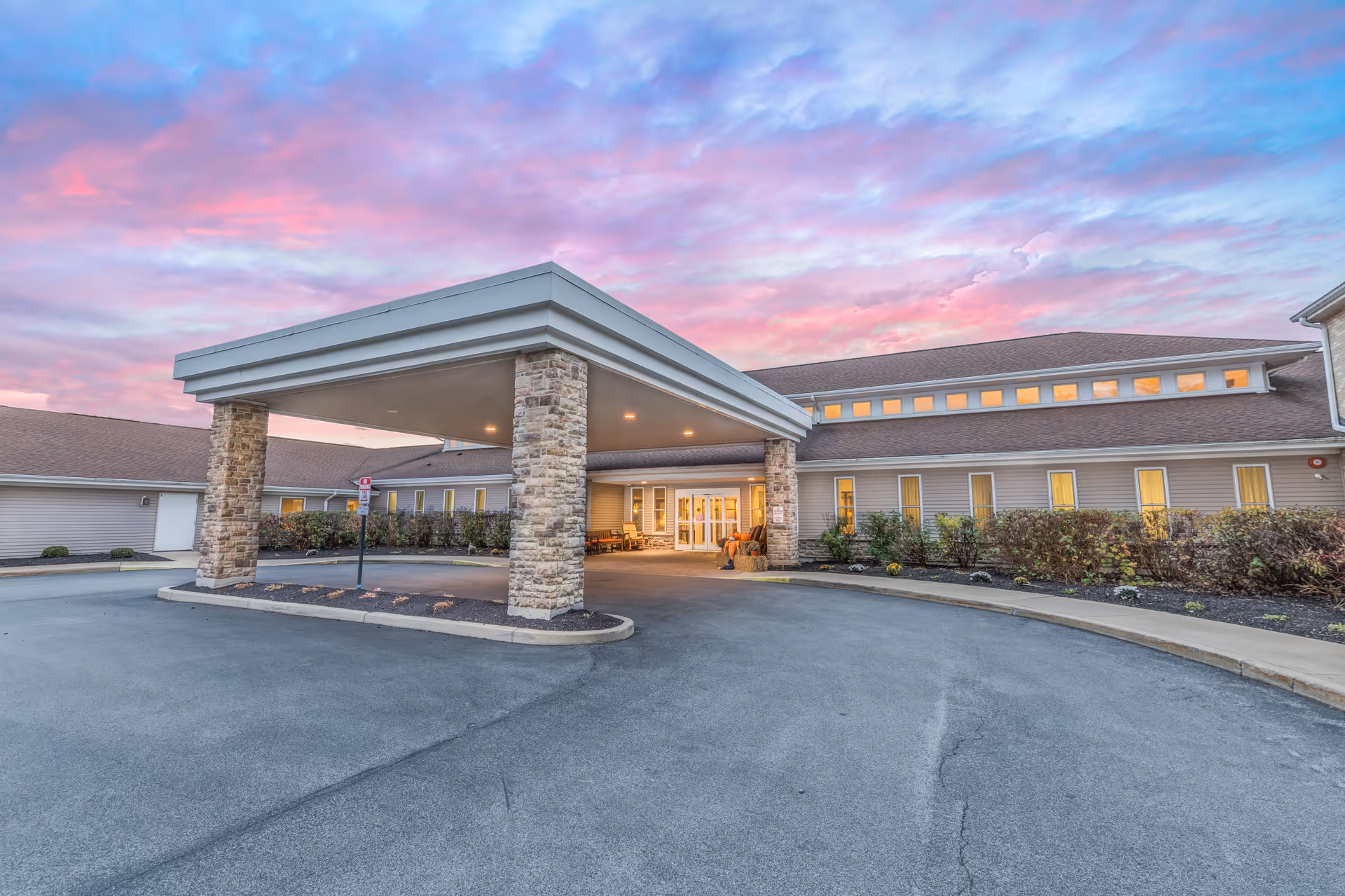 Exterior view of Brompton Heights of Williamsville facility entrance at dusk, featuring a covered drop-off area supported by stone pillars, surrounded by a paved driveway and landscaped bushes, under a colorful pink and blue sky.