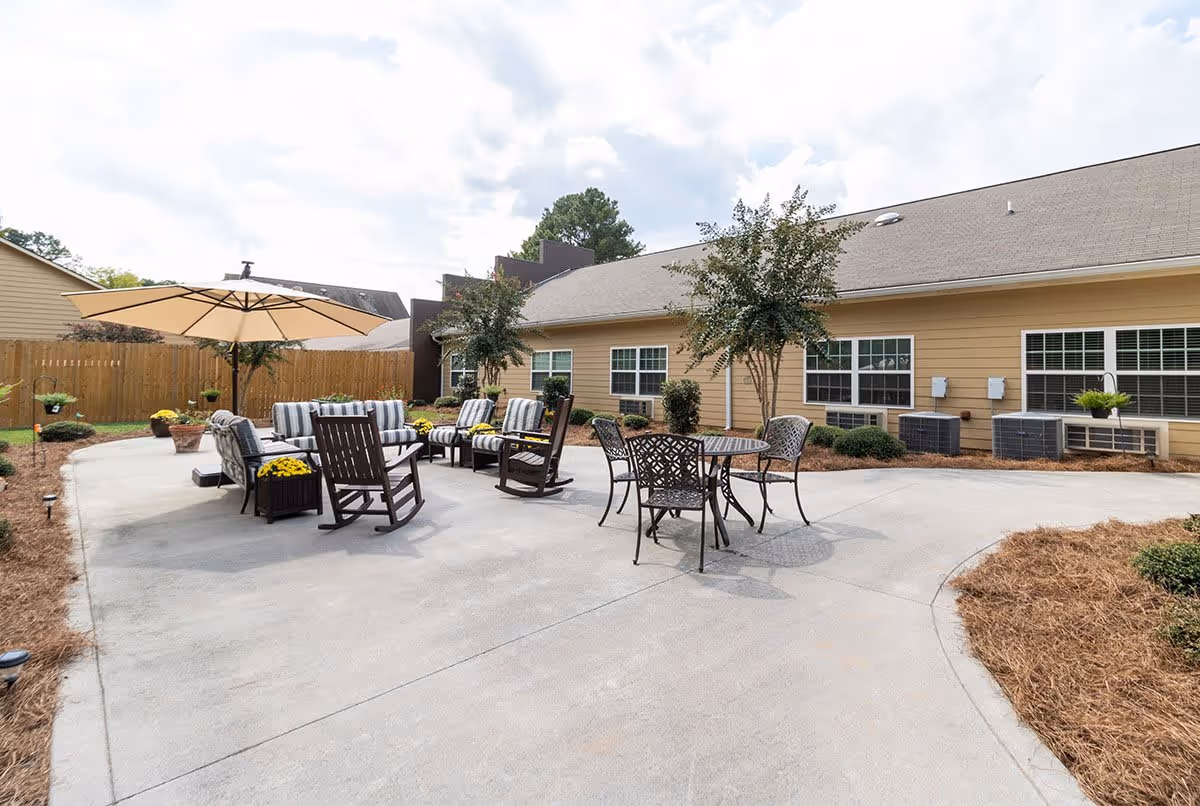 Outdoor patio area with multiple seating options including cushioned chairs, rocking chairs, and a metal table with chairs. There is a large beige umbrella providing shade over some of the seating. The patio is surrounded by a wooden fence, small trees, and landscaped mulch beds. The building in the background has beige siding and several windows.