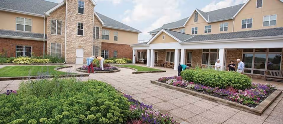 Outdoor courtyard area of a senior living facility with well-maintained flower beds and shrubs. Several elderly people are tending to the garden and socializing near the building, which has a brick and stone exterior with large windows and a covered patio.