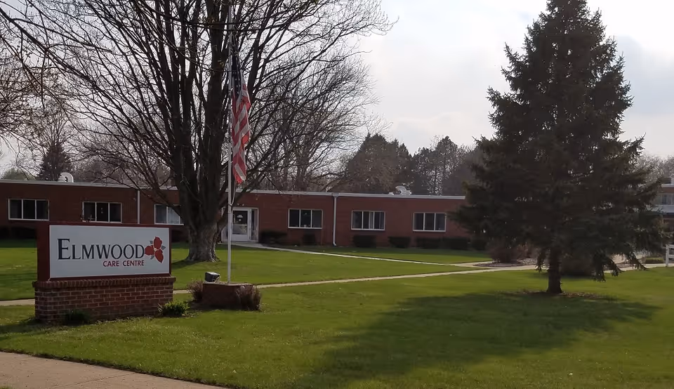 Low single-story brick care facility with a lawn, flagpole, trees, and a sign reading "Elmwood Care Centre".