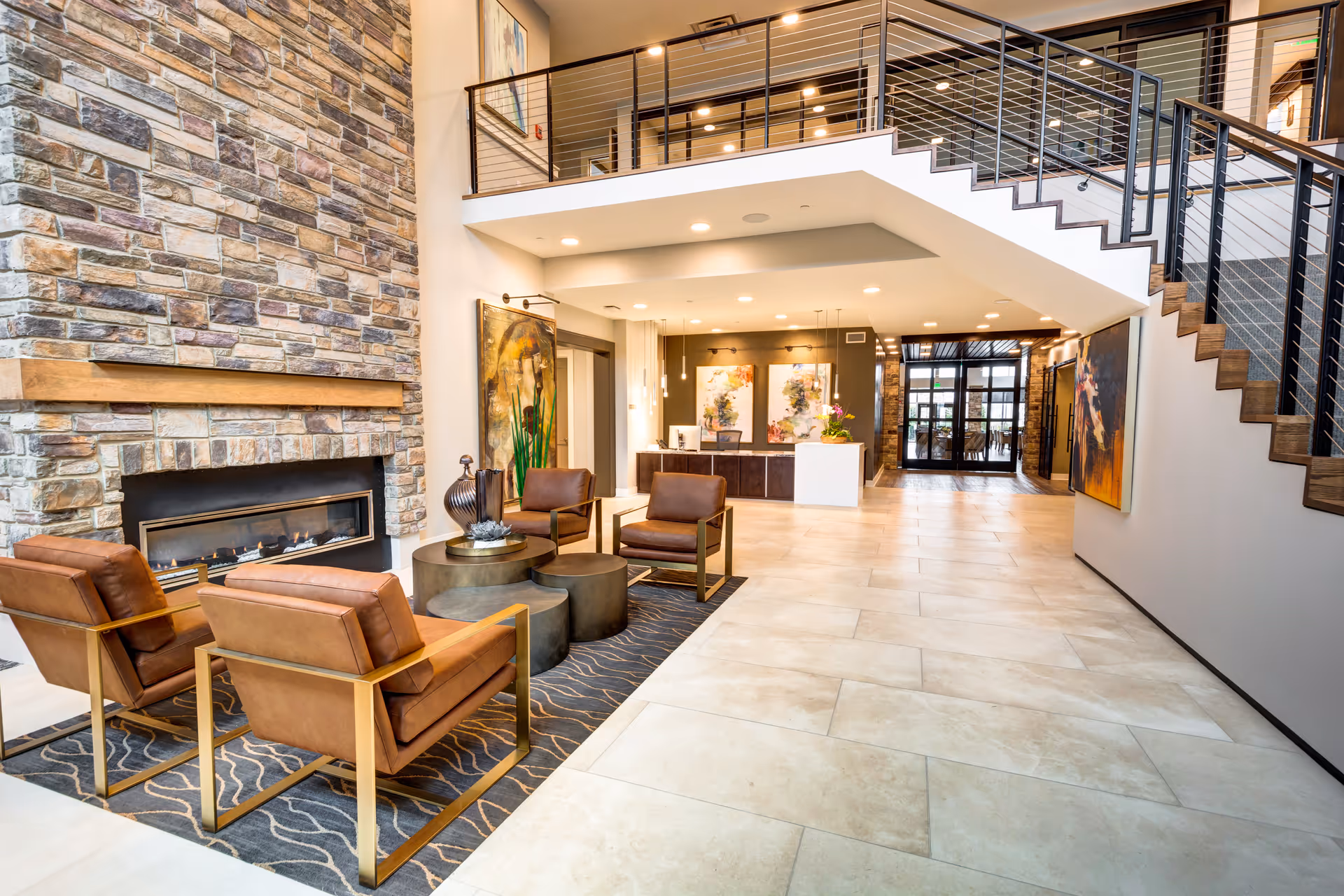 A spacious and modern senior living facility lobby with a stone fireplace, four brown leather chairs arranged around a set of round coffee tables on a patterned rug, a reception desk in the background, and a staircase with metal railings leading to an upper floor.