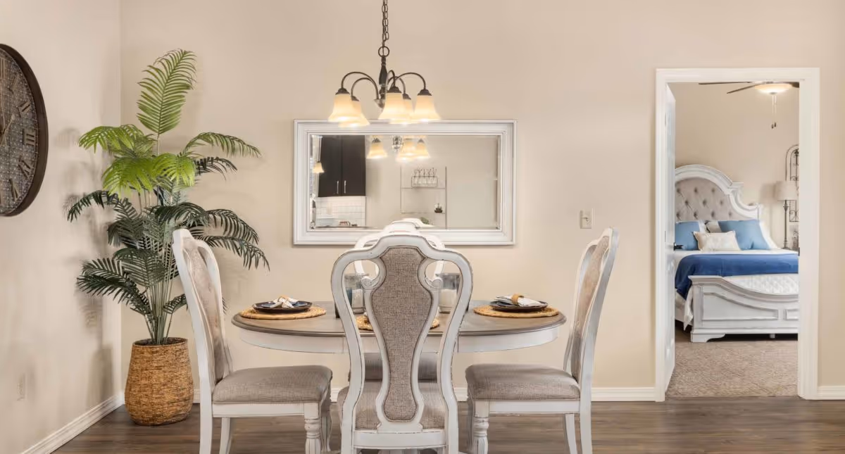 Dining area with a round table and upholstered chairs, a potted plant, wall mirror, and a bedroom visible through a doorway.