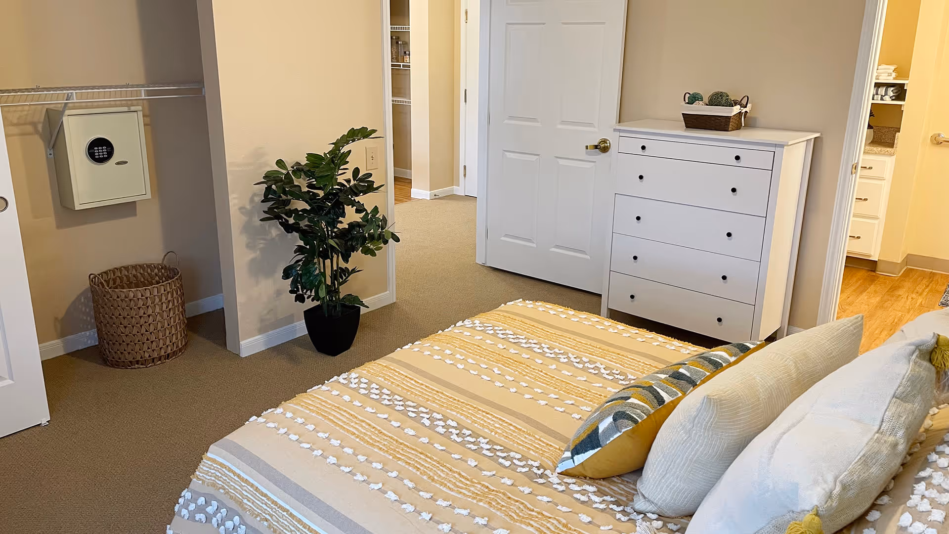 A bedroom with a bed covered in a beige and white textured blanket and several pillows. There is a white dresser with a basket on top, a green potted plant, an open closet with a safe mounted on the wall, and a view into an adjacent bathroom with wooden flooring.