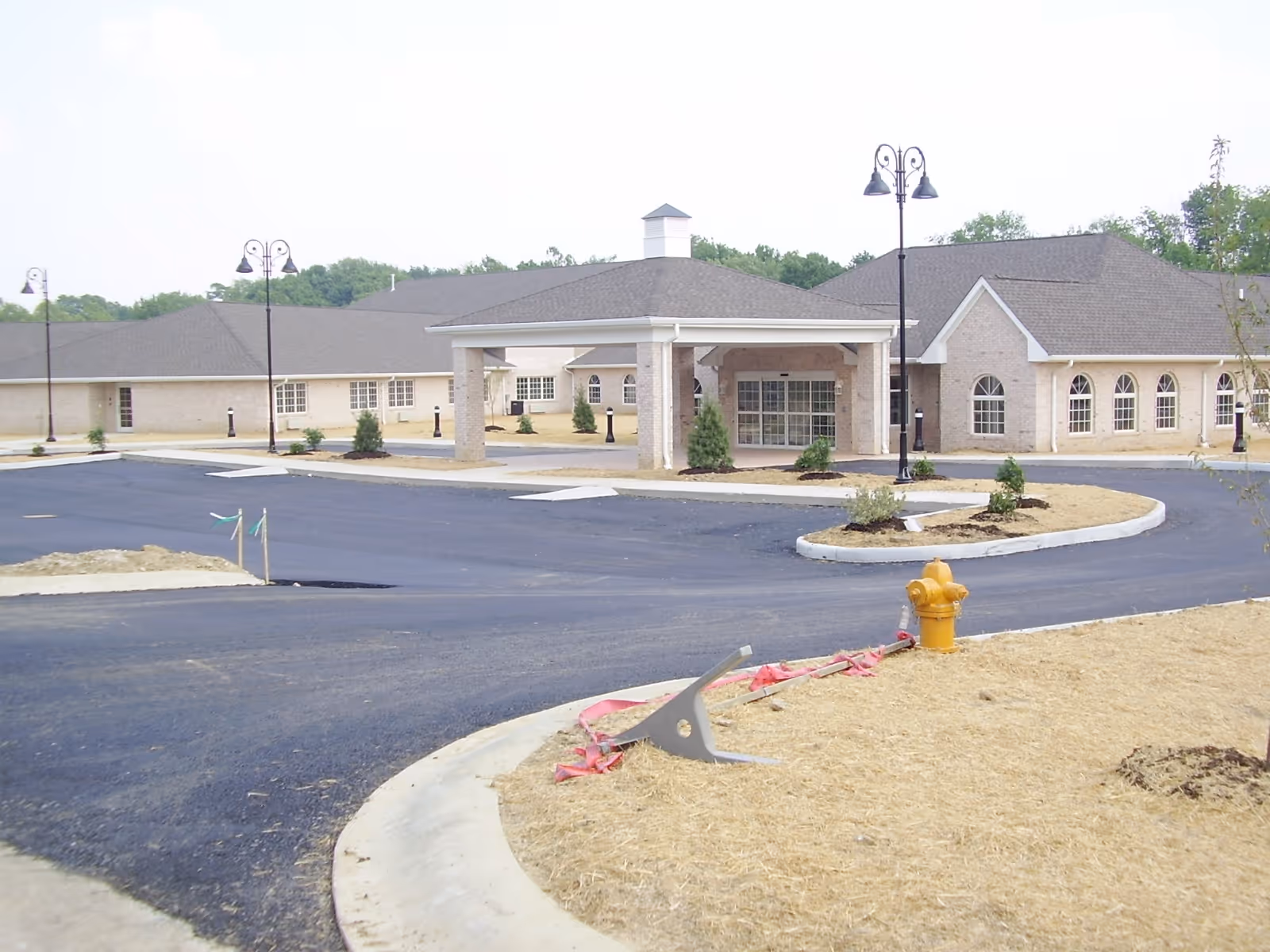 Exterior view of a single-story senior living facility building with a covered entrance, newly paved driveway, street lamps, small landscaped areas, and a yellow fire hydrant in the foreground.