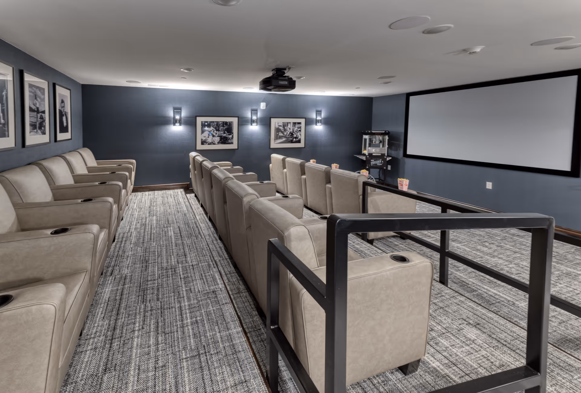 A cozy senior living facility movie theater room with two rows of beige reclining chairs facing a large blank projection screen. The room has dark blue walls adorned with black and white framed photographs and wall sconces providing soft lighting. A popcorn machine is visible in the corner, and the floor is carpeted with a patterned gray carpet.