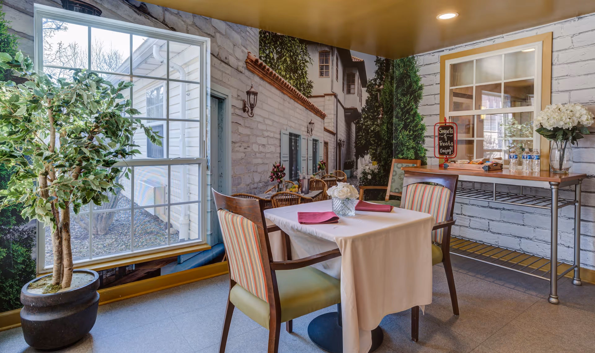 A small dining area with a table covered by a white tablecloth and two chairs with striped upholstery. On the table is a vase with white flowers and two folded red napkins. Behind the table is a metal cart with bottled water, a vase of white flowers, and a small chalkboard sign that reads 'Sweets & Treats Tonight!'. The walls have a mural depicting an outdoor scene with a stone building, greenery, and a window. A large potted plant is placed near the window mural.