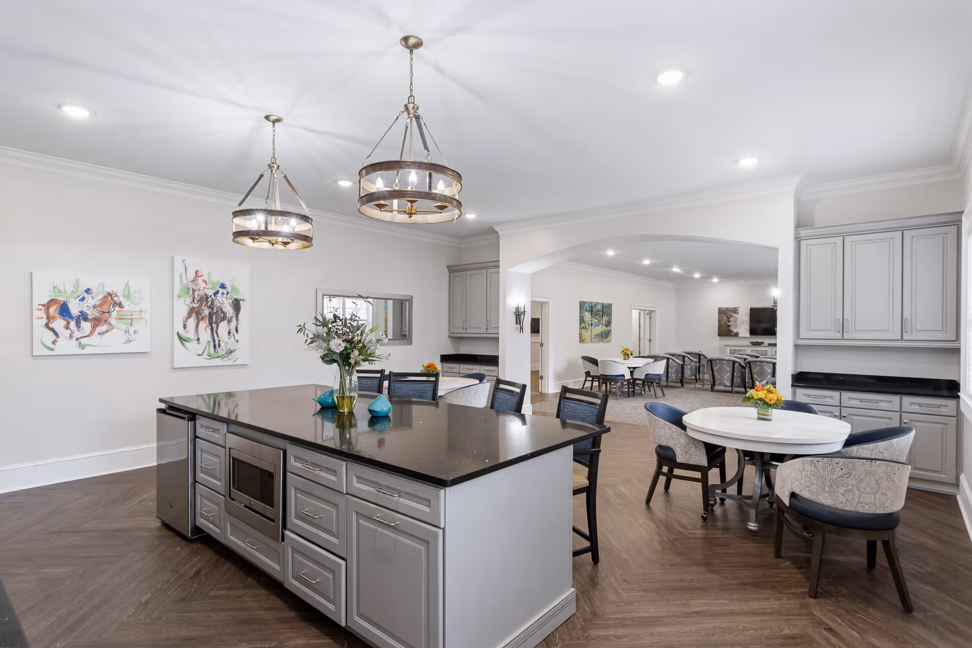 A spacious, modern kitchen and dining area in a senior living facility. The kitchen features a large island with a black countertop, built-in microwave, and multiple drawers and cabinets. Two pendant lights hang above the island. There are round dining tables with upholstered chairs, floral centerpieces, and artwork on the walls. The room has wood flooring and neutral-colored walls with recessed lighting.