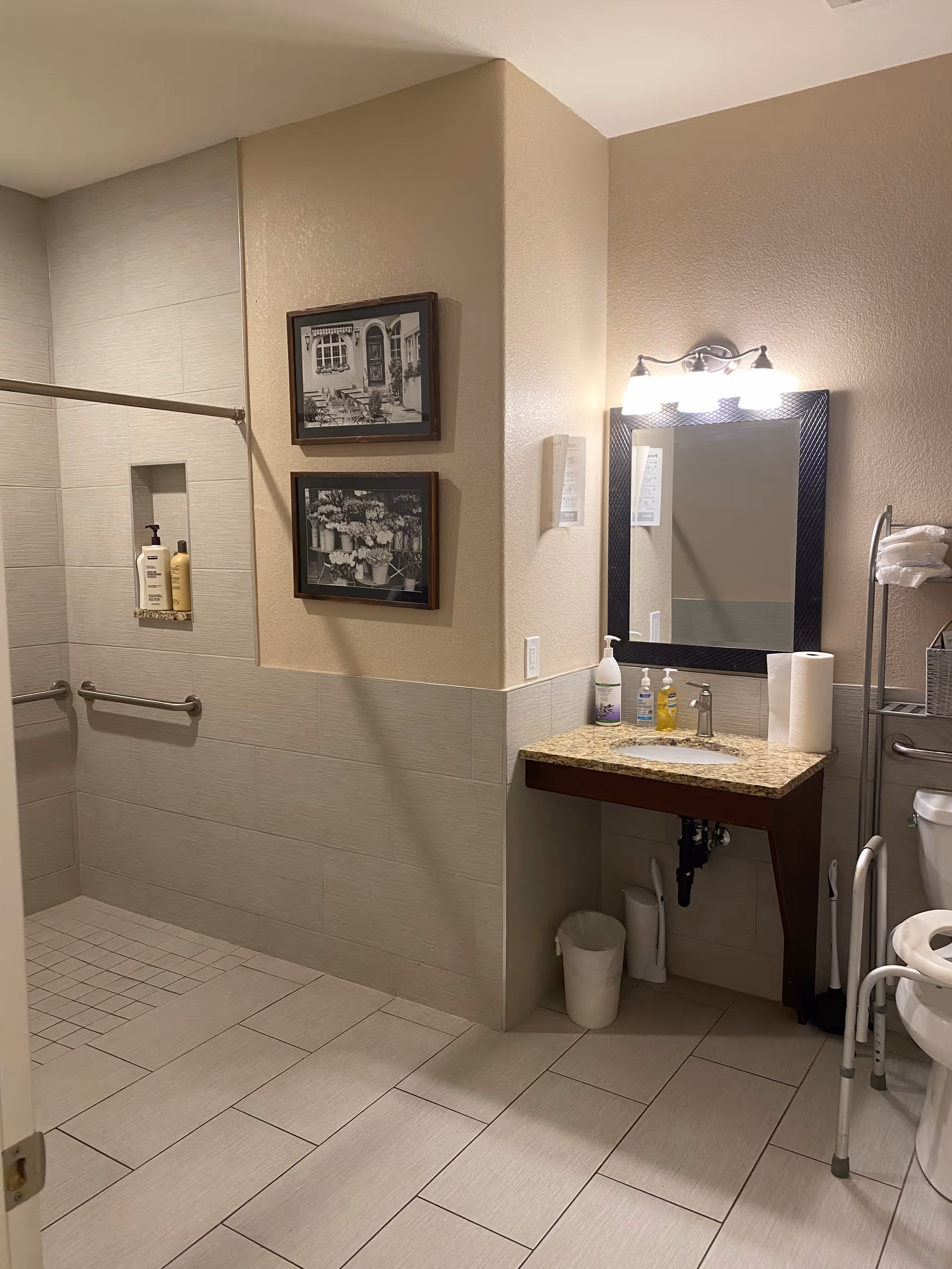 A clean, accessible bathroom with beige tiled walls and floor. The room features a walk-in shower with grab bars and shampoo bottles, a granite countertop sink with a mirror and light fixture above, a toilet with a safety frame, and a metal rack holding towels and a basket. Two black and white framed pictures hang on the wall near the shower.