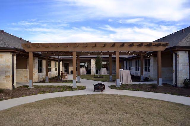 Outdoor courtyard area of a senior living facility with a wooden pergola structure, stone buildings surrounding the courtyard, a fire pit in the center, and several tall cocktail tables covered with beige cloth. The sky is partly cloudy.