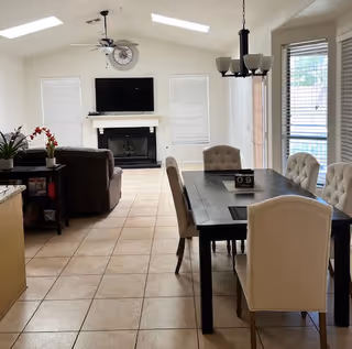 A bright and spacious living and dining area in an assisted living facility. The room features a tiled floor, a dark wooden dining table with six cushioned chairs, a chandelier above the table, and a large flat-screen TV mounted above a white fireplace. There are windows with blinds on the walls, a ceiling fan, and a small table with plants and decorative items near a brown sofa.