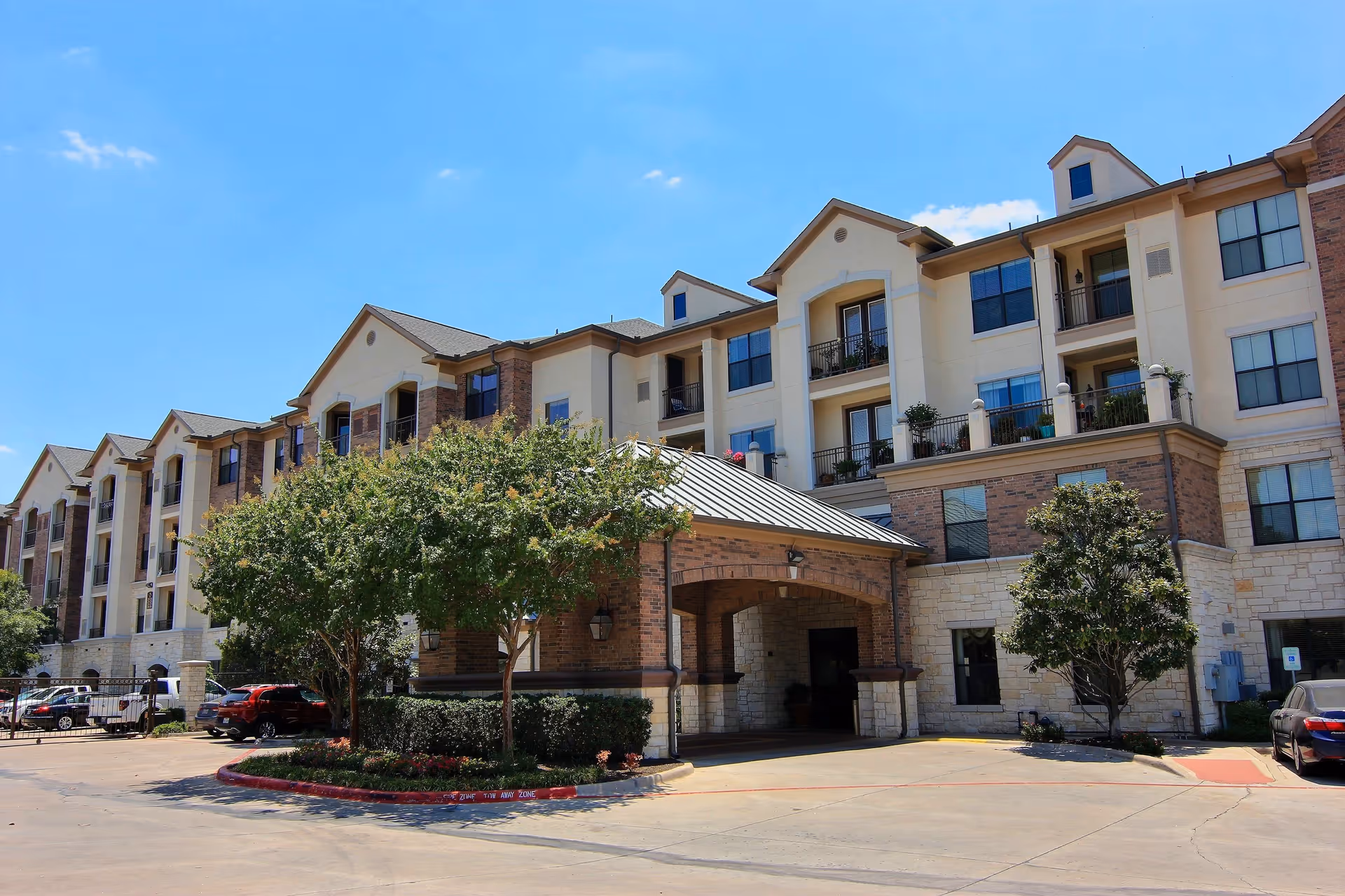 Exterior view of a multi-story senior living facility building with balconies, trees, and parked cars under a clear blue sky.