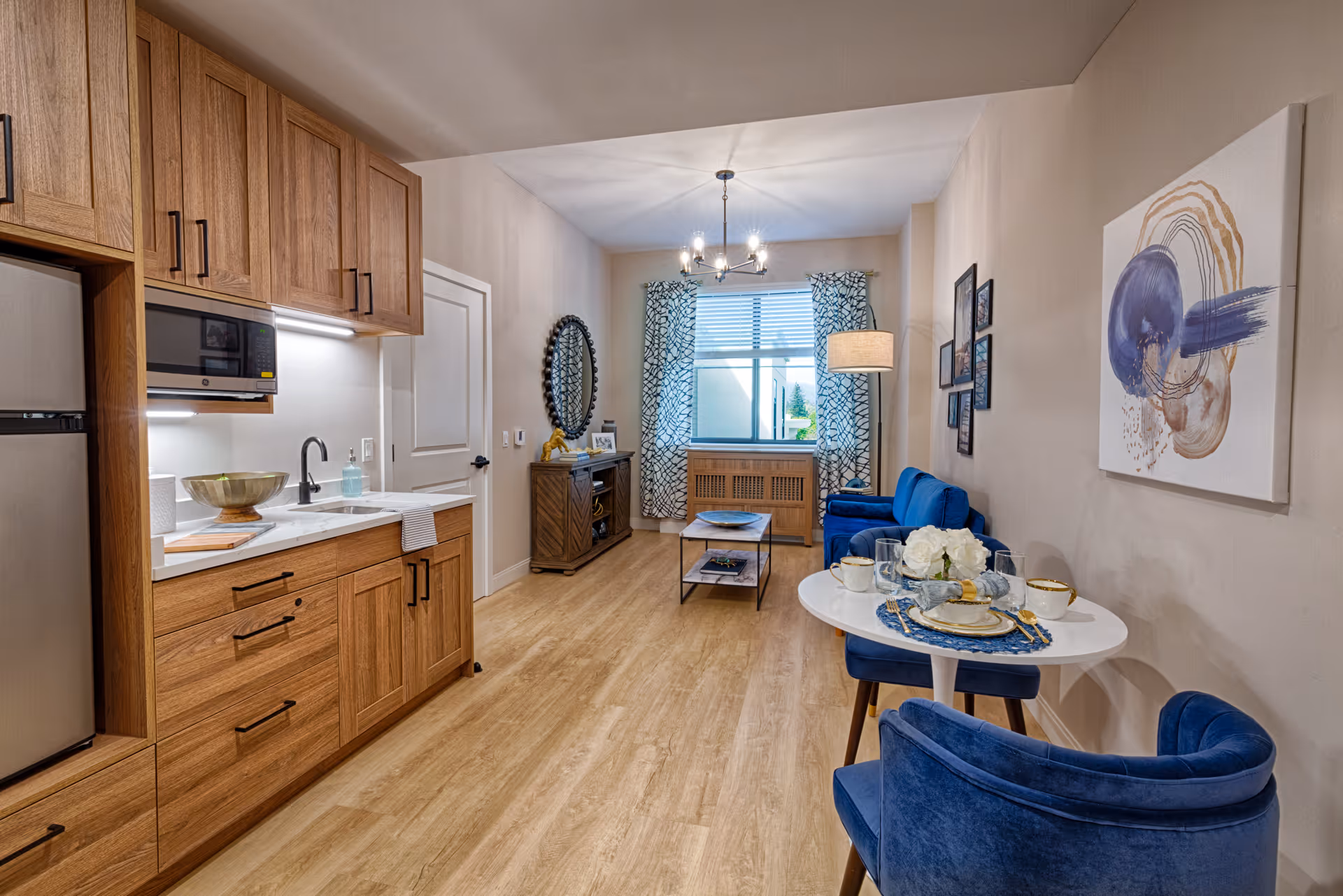 Interior view of a senior living facility apartment featuring a small kitchen area with wooden cabinets, a microwave, and a sink on the left. The room extends into a living area with a blue sofa, a coffee table, a floor lamp, and a window with patterned curtains. In the foreground, there is a small round dining table set with plates, cups, and a flower centerpiece, surrounded by blue upholstered chairs. The walls are decorated with framed pictures and abstract artwork.