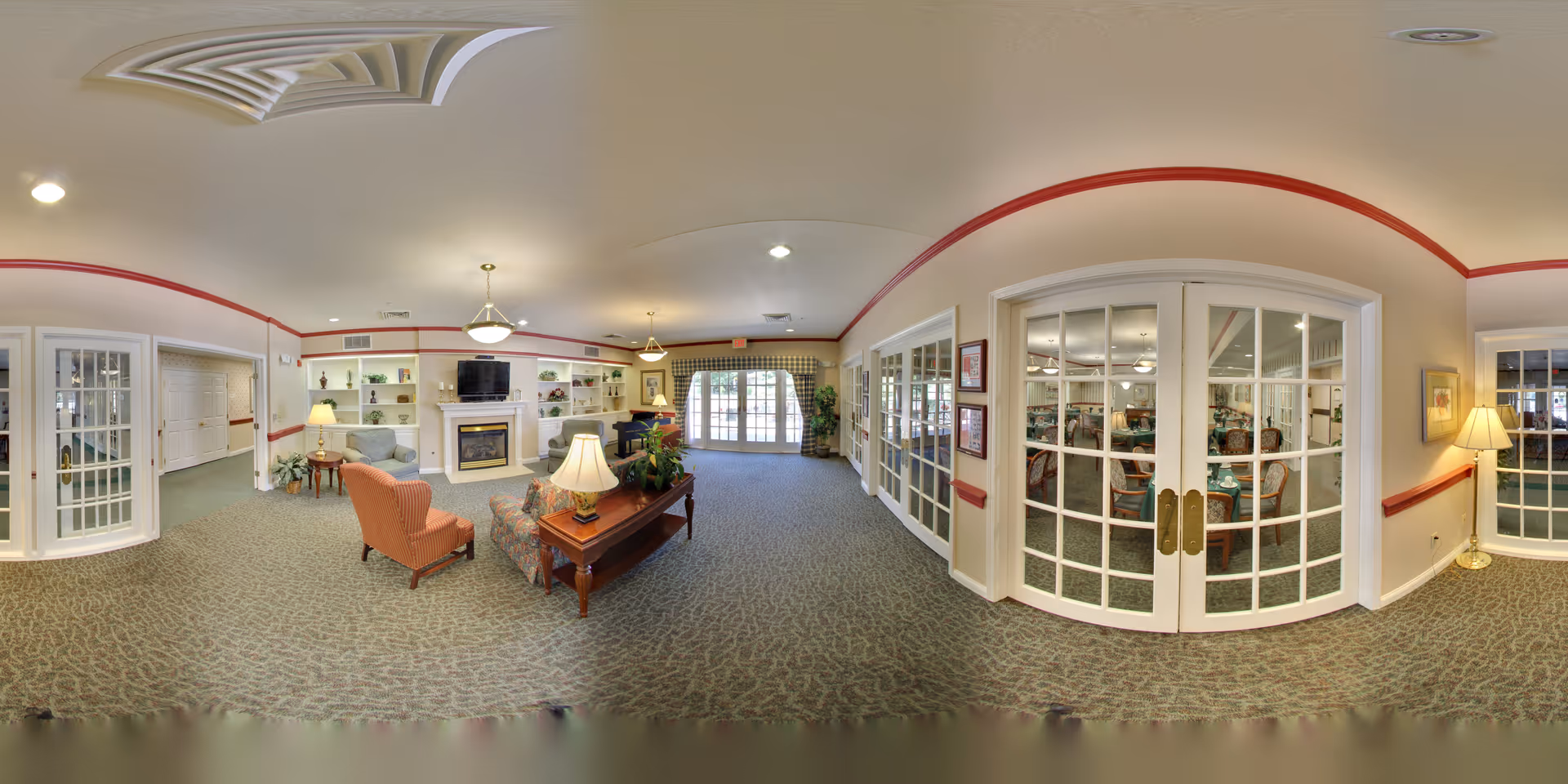 A spacious and well-lit common area in a senior living facility featuring comfortable seating including armchairs and a sofa around a wooden coffee table with a lamp. The room has a fireplace with a TV mounted above it, built-in shelves with decorative items, and large glass doors leading to another room with tables and chairs. The walls are cream-colored with red trim, and the carpet has a patterned design. There are ceiling lights and lamps providing warm lighting throughout the space.