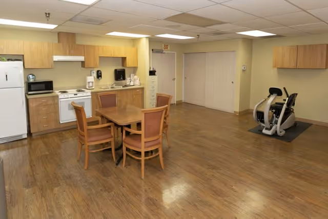Multipurpose common room with a small kitchenette, a wooden dining table and four chairs, and an exercise bike on a wood floor.