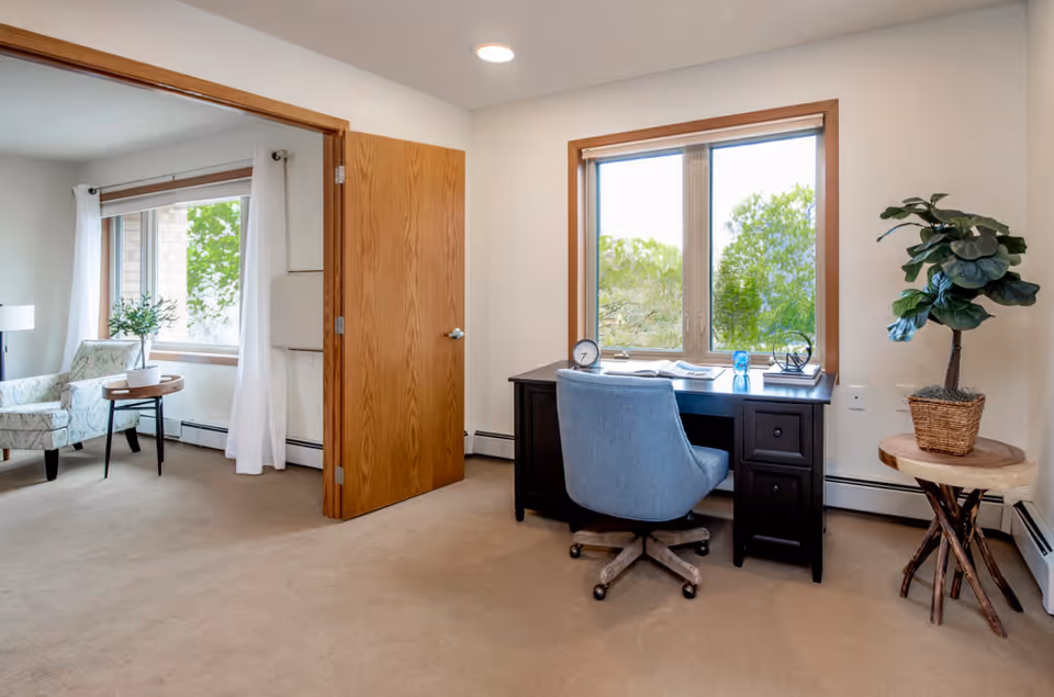 Bright interior room with a desk and chair by a window and a seating area with an armchair visible through an open door.
