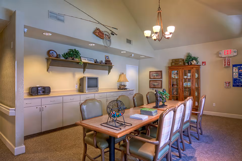 A well-lit room with a long wooden table surrounded by eight chairs, some with green cushions and others with plaid upholstery. On the table are a bingo cage with colorful balls, a stack of cards, and a small statue. The room has beige walls, a carpeted floor, and a chandelier hanging from the ceiling. Against one wall is a long counter with cabinets underneath, a small TV, a radio, and decorative items including a 'Gone Fishing' sign and fishing gear mounted above. A wooden cabinet with glass doors displaying plates and other items is in the corner, along with framed pictures on the wall and an exit sign above a door.