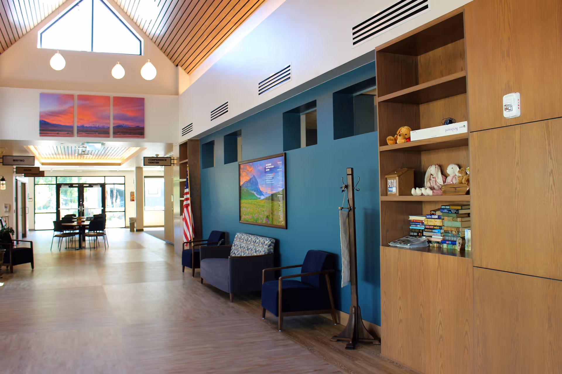 Bright senior living facility lobby with seating, a bookshelf, an American flag, and a high vaulted ceiling with pendant lights.