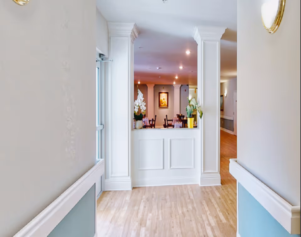 View down a hallway with light wooden flooring and white walls with blue paneling. At the end of the hallway is a white counter with decorative columns on either side, topped with potted plants. Beyond the counter is a dining area with tables and chairs, soft lighting, and a framed picture on the wall.