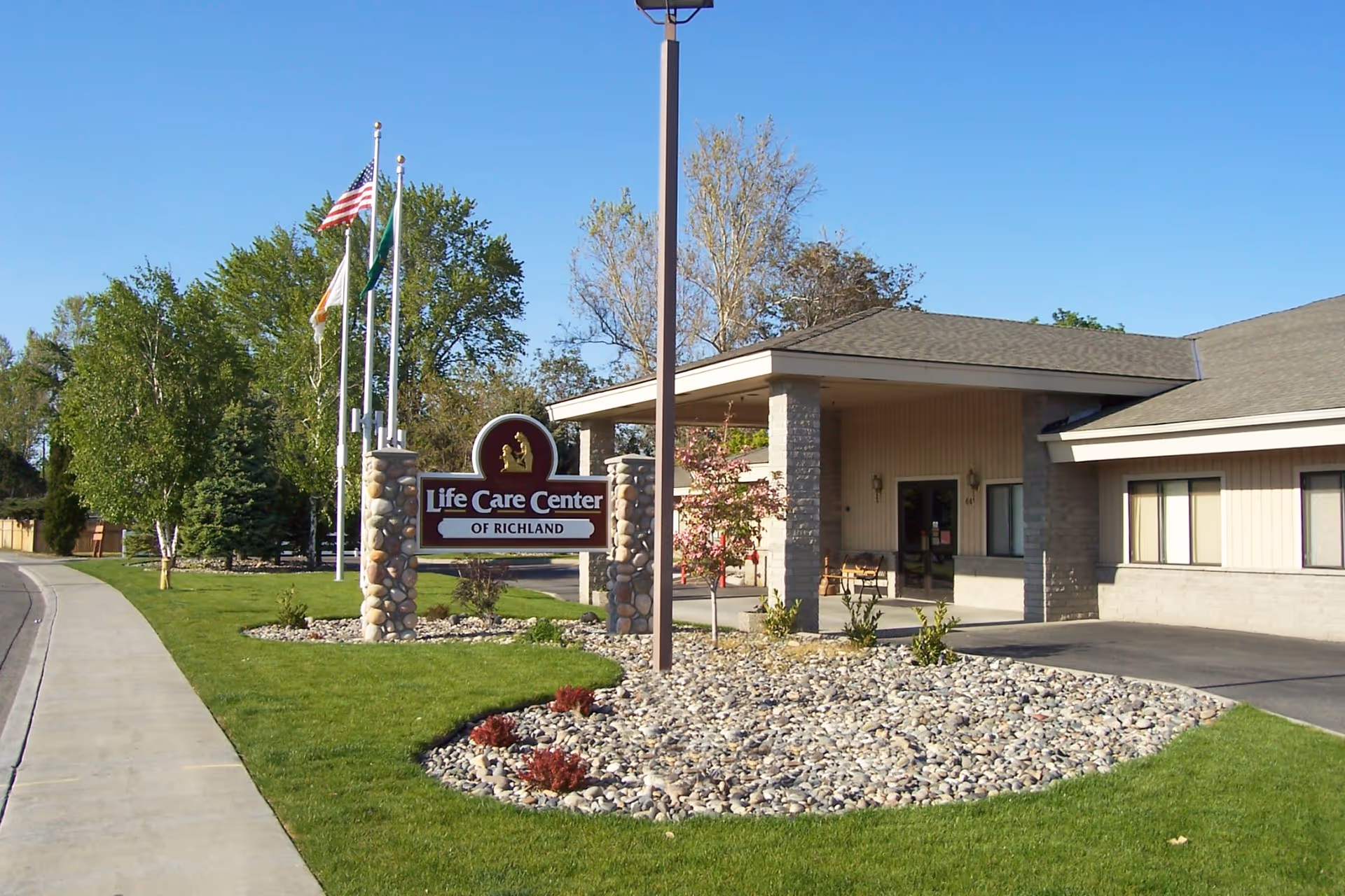 Exterior view of the Life Care Center of Richland building with a stone sign in front, surrounded by green grass, small plants, and trees. Three flagpoles with flags are visible near the sign, and a sidewalk runs along the left side of the image under a clear blue sky.