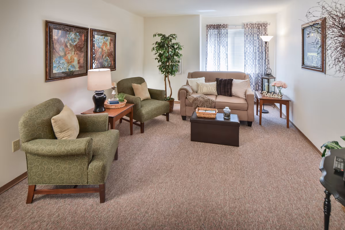 A cozy living room with a beige loveseat adorned with pillows and a throw blanket, two green patterned armchairs with beige cushions, a wooden side table with a lamp and decorative items, a small coffee table with a basket and candle, a tall floor lamp, and framed floral artwork on the walls. The room has carpeted flooring and a window with patterned curtains letting in natural light.