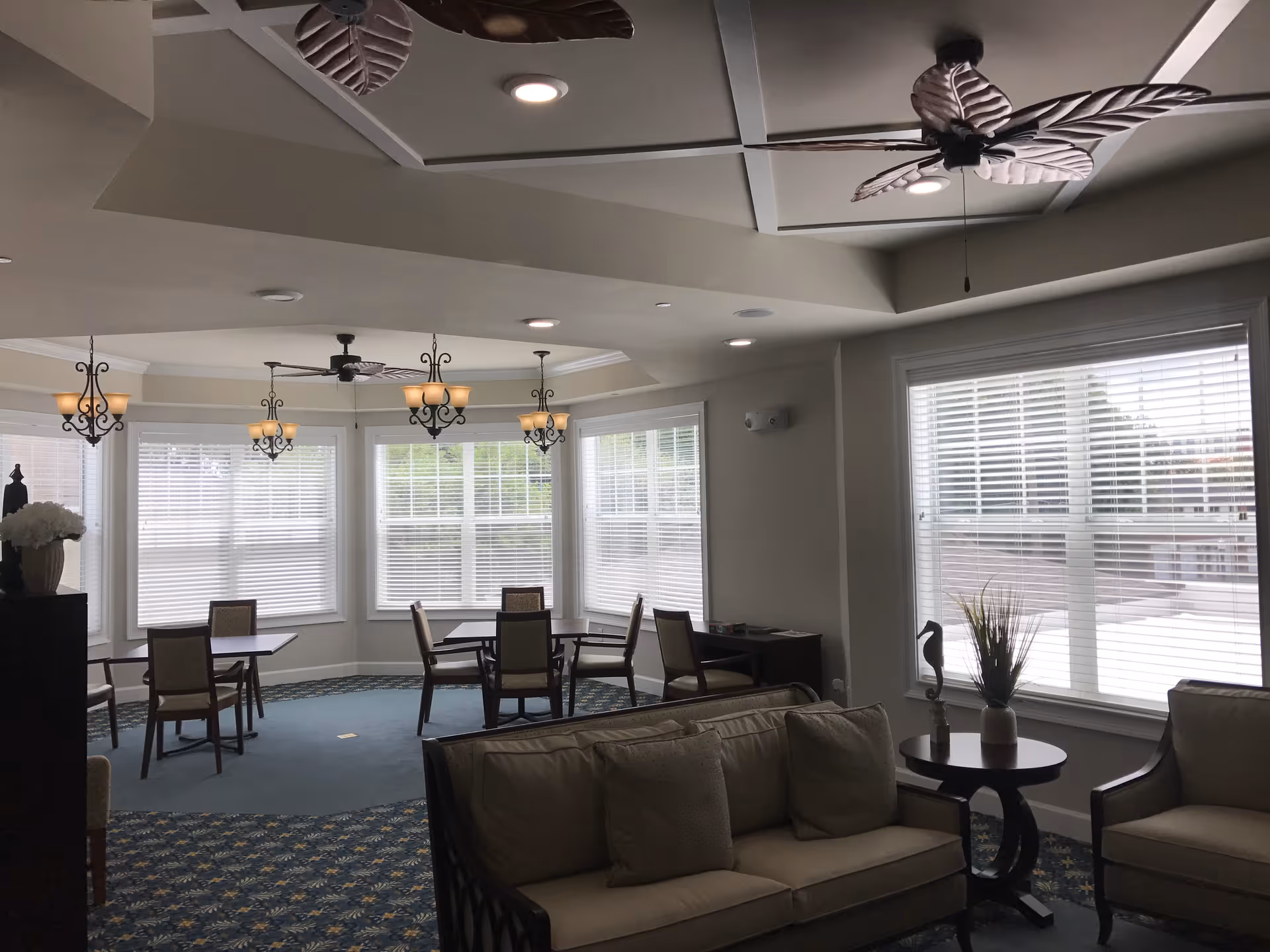 A bright and spacious common area with multiple windows covered by white blinds. The room features several tables with chairs, a sofa with cushions, an armchair, and a small round table with a plant and decorative item. The ceiling has recessed lighting, ceiling fans with leaf-shaped blades, and hanging light fixtures.