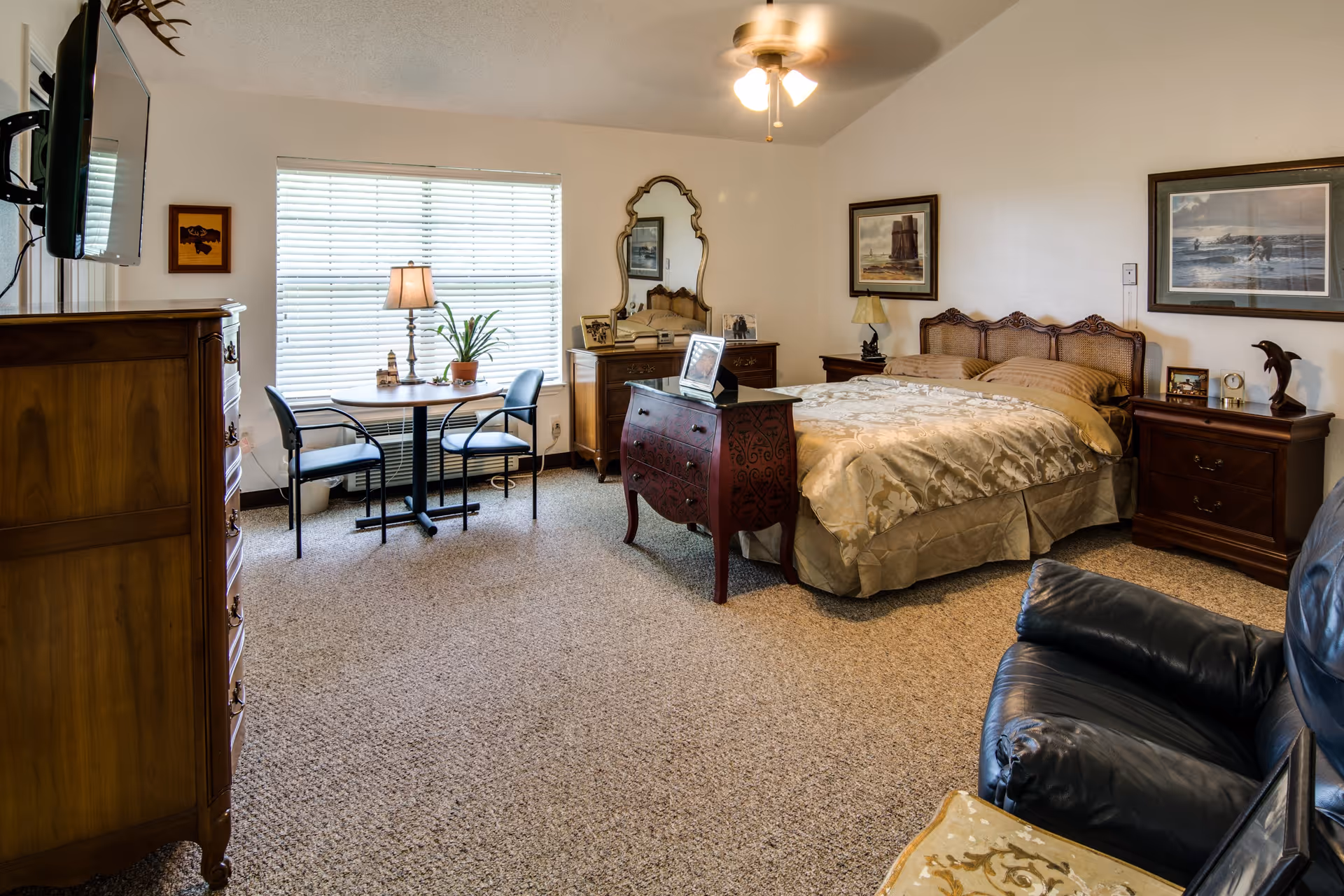 A cozy bedroom with a large bed covered in a beige patterned comforter, two wooden nightstands with lamps and decorative items, a wooden dresser with a mirror, a small table with two chairs near a window with blinds, a mounted TV on the wall, and a dark leather armchair in the foreground.