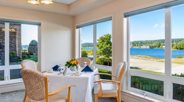 A dining area with a table set for four, featuring a white tablecloth, blue napkins, and a floral centerpiece. The room has large windows offering a scenic view of a lake and greenery outside.