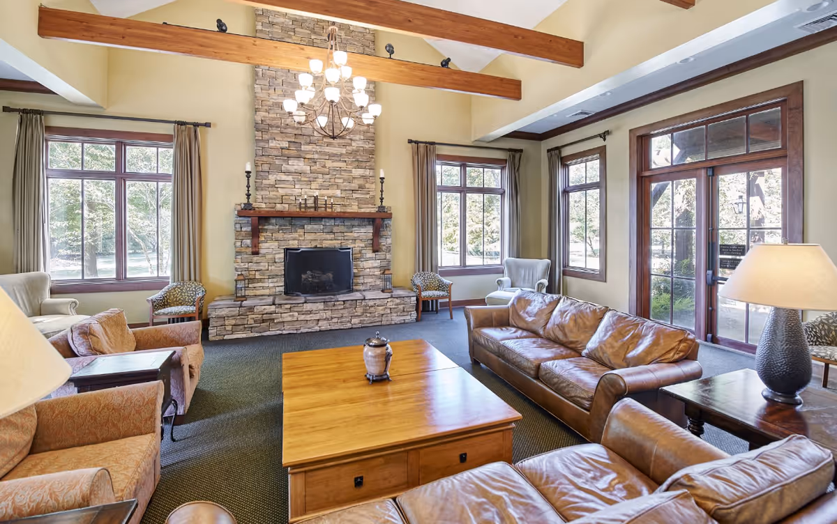 A spacious living room with a stone fireplace centered on the far wall, flanked by large windows with beige curtains. The room features wooden ceiling beams, a chandelier, leather sofas, armchairs, a wooden coffee table, and side tables with lamps. Glass doors lead outside, allowing natural light to fill the room.