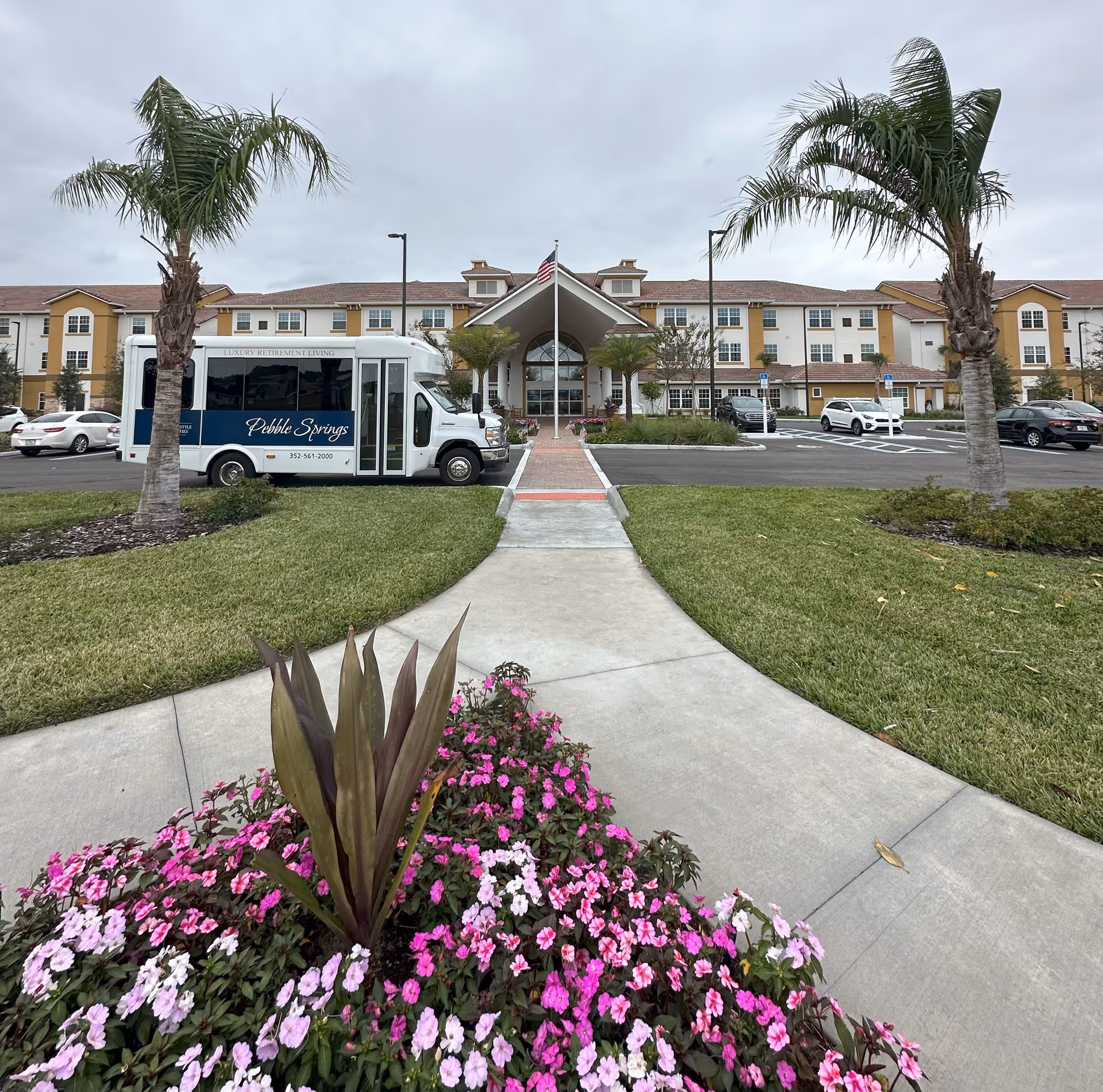 Front exterior view of Pebble Springs Retirement Community building with a pathway leading to the entrance, flanked by palm trees and flower beds. A white and blue shuttle bus with the Pebble Springs logo is parked in front, along with several cars in the parking lot under a cloudy sky.