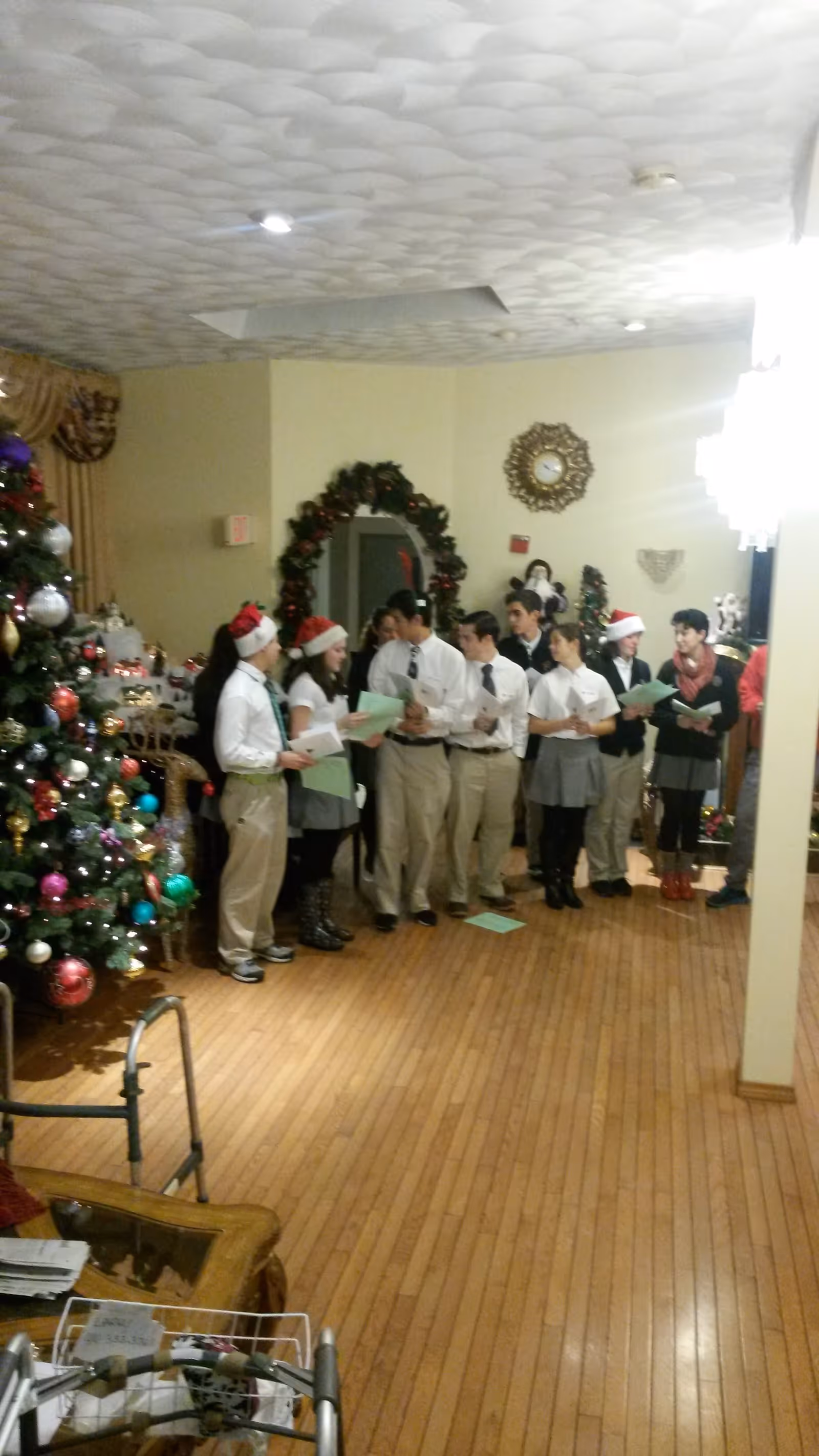 A group of people, some wearing Santa hats, standing in a line inside a decorated room with a Christmas tree and holiday wreaths. They appear to be singing or reading from sheets of paper. The room has wooden floors and festive decorations.