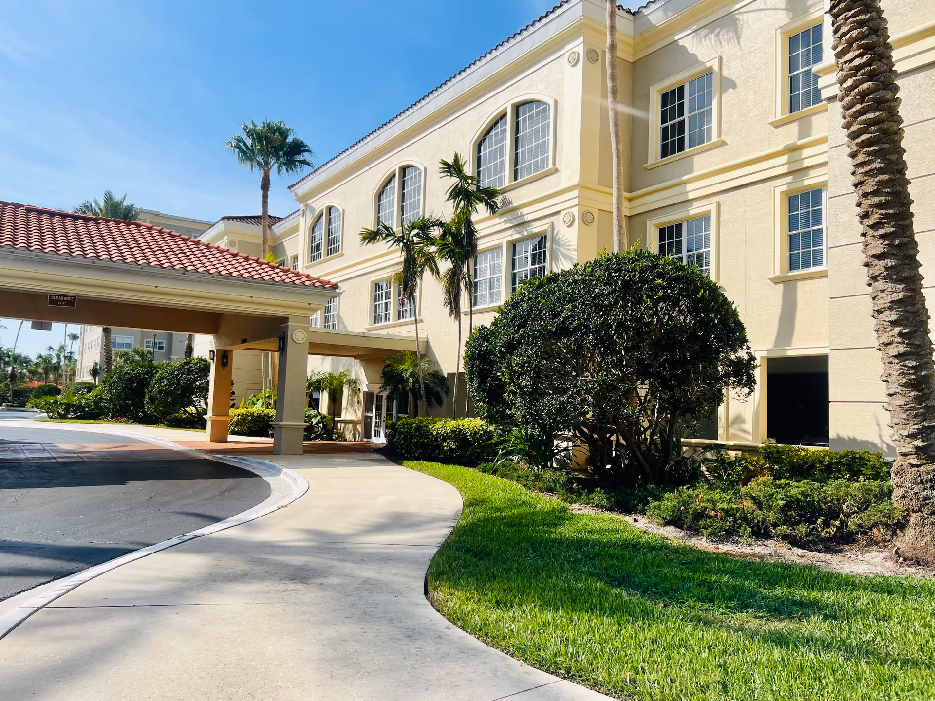 Front entrance of a yellow Mediterranean-style senior living building with a covered porte-cochere, palm trees, and landscaped grounds.