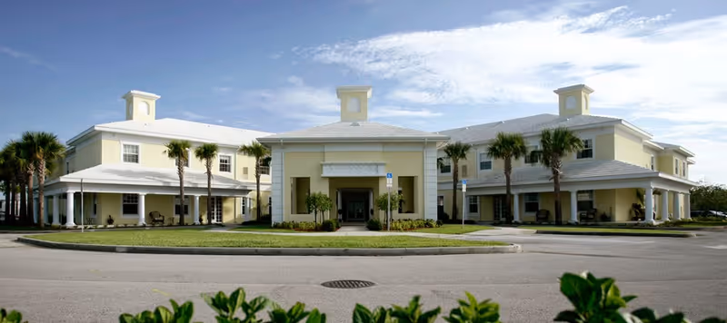 Front exterior view of a two-story assisted living facility building with light yellow walls, white roof, and multiple palm trees around the entrance. The building has a covered porch area and a circular driveway in front.