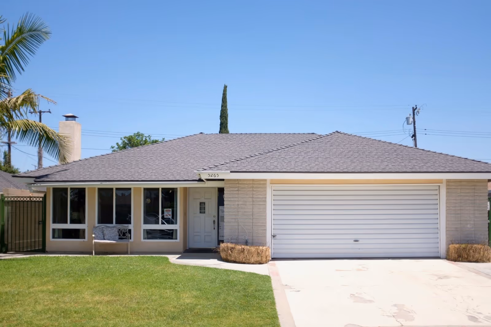 Front exterior view of a single-story house with a gray shingled roof, beige walls, a white garage door, and a white front door. There is a green lawn in front, a palm tree on the left, and a bench near the windows. The house number 5265 is visible above the front door.