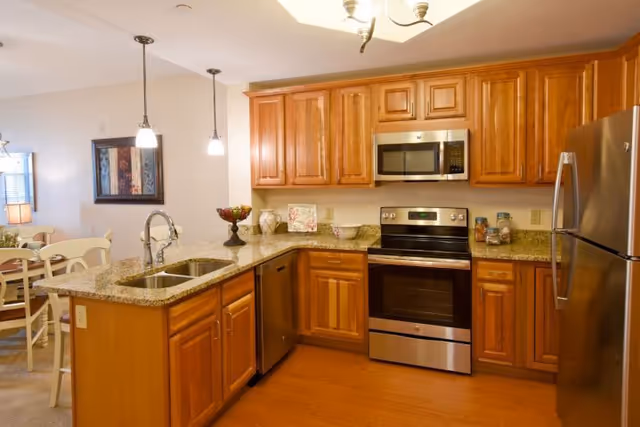 A modern kitchen with wooden cabinets, granite countertops, stainless steel appliances including a refrigerator, oven, microwave, and dishwasher. There is a double sink on the island with two pendant lights hanging above. In the background, a dining area with a table and chairs is partially visible.