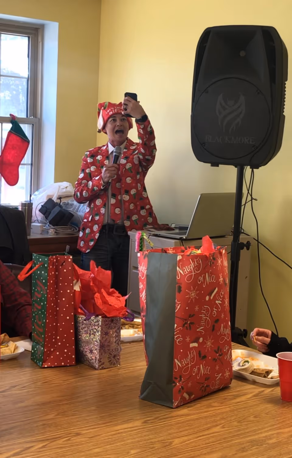 A person wearing a festive red jacket with snowman patterns and a matching Santa hat is holding a microphone and taking a selfie in a room decorated for Christmas. There are several gift bags with holiday designs on a wooden table in the foreground, and a Christmas stocking hanging by a window in the background. A large speaker and a laptop are also visible behind the person.