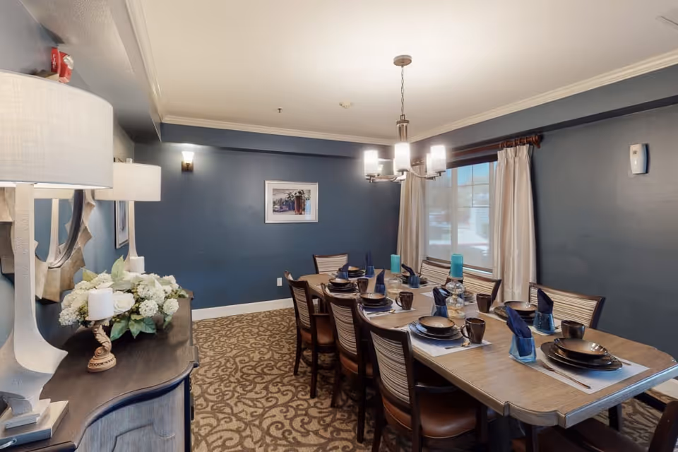 A dining room with a long wooden table set for eight people with brown plates, mugs, and blue napkins. The room has blue walls, a patterned carpet, a window with beige curtains, a chandelier overhead, and a sideboard with a lamp, candles, and a floral arrangement.